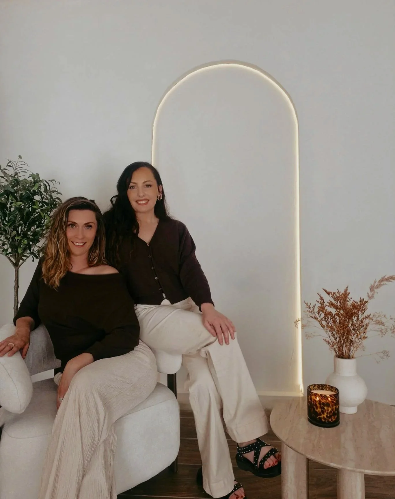 Two women sitting on a light-colored sofa in a modern room with a tall white wall, a potted plant, a small table with a vases and a candle, both smiling and looking at the camera.