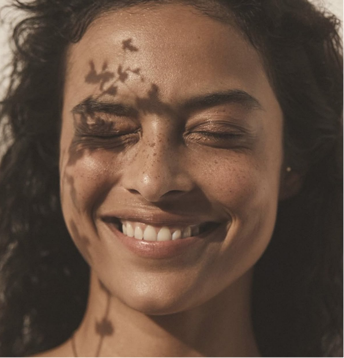 Close-up of a smiling woman with curly hair and skin freckles, with a shadow of leaves on her face and neck.