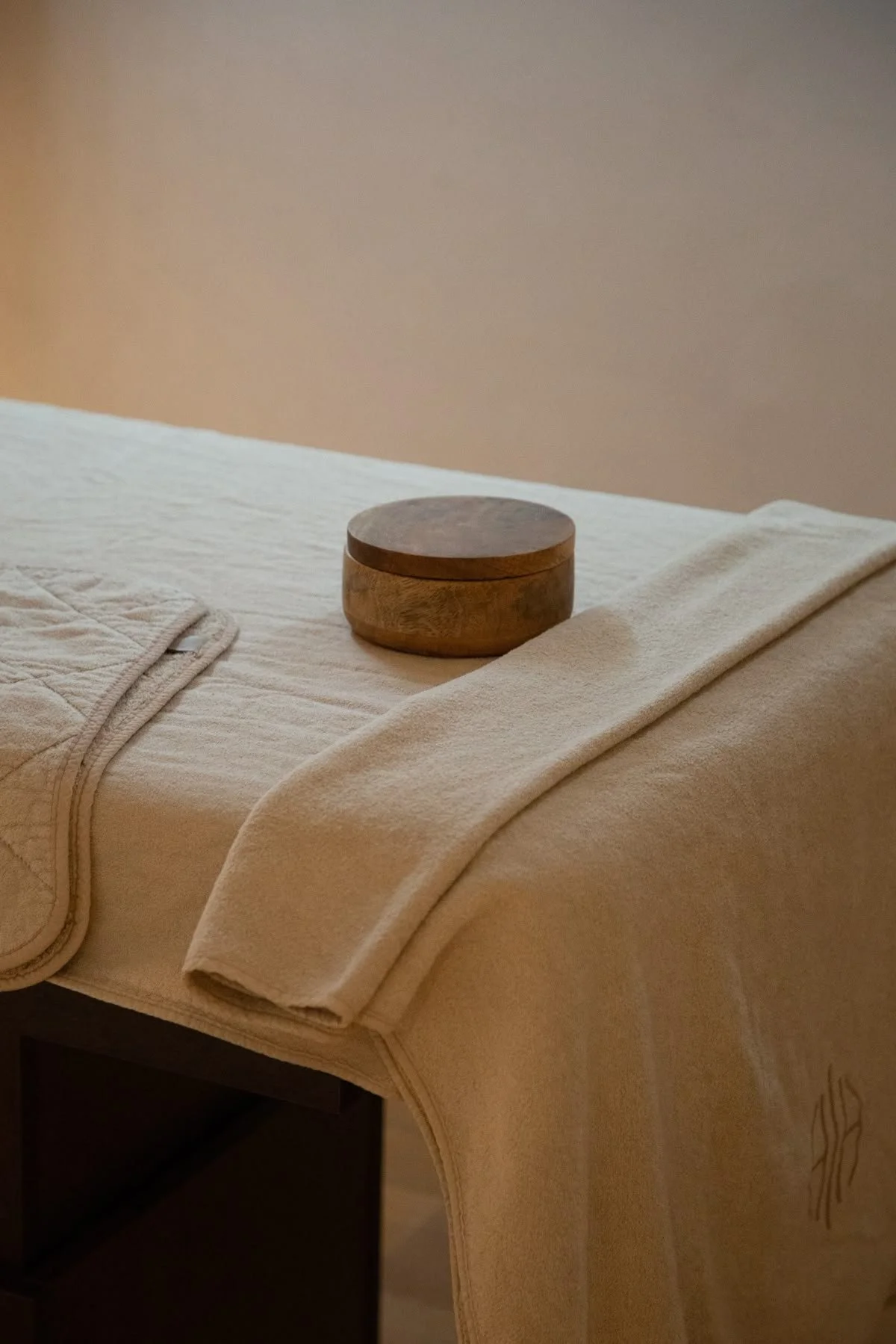 A neatly made massage table covered with a cream-colored towel, with a small round wooden container placed on top of the towel.