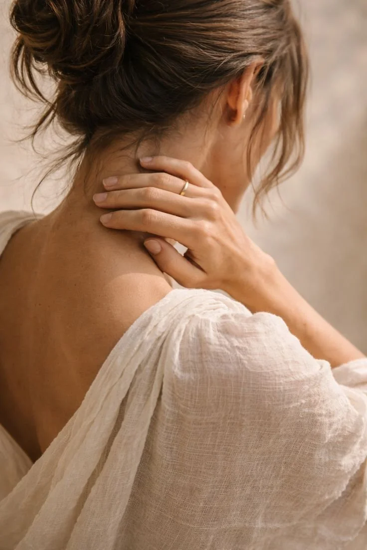 A woman with light brown hair, tied back in a loose bun, is touching her neck and shoulder with her hand. She is wearing a light, textured fabric top, and the background is neutral and out of focus.