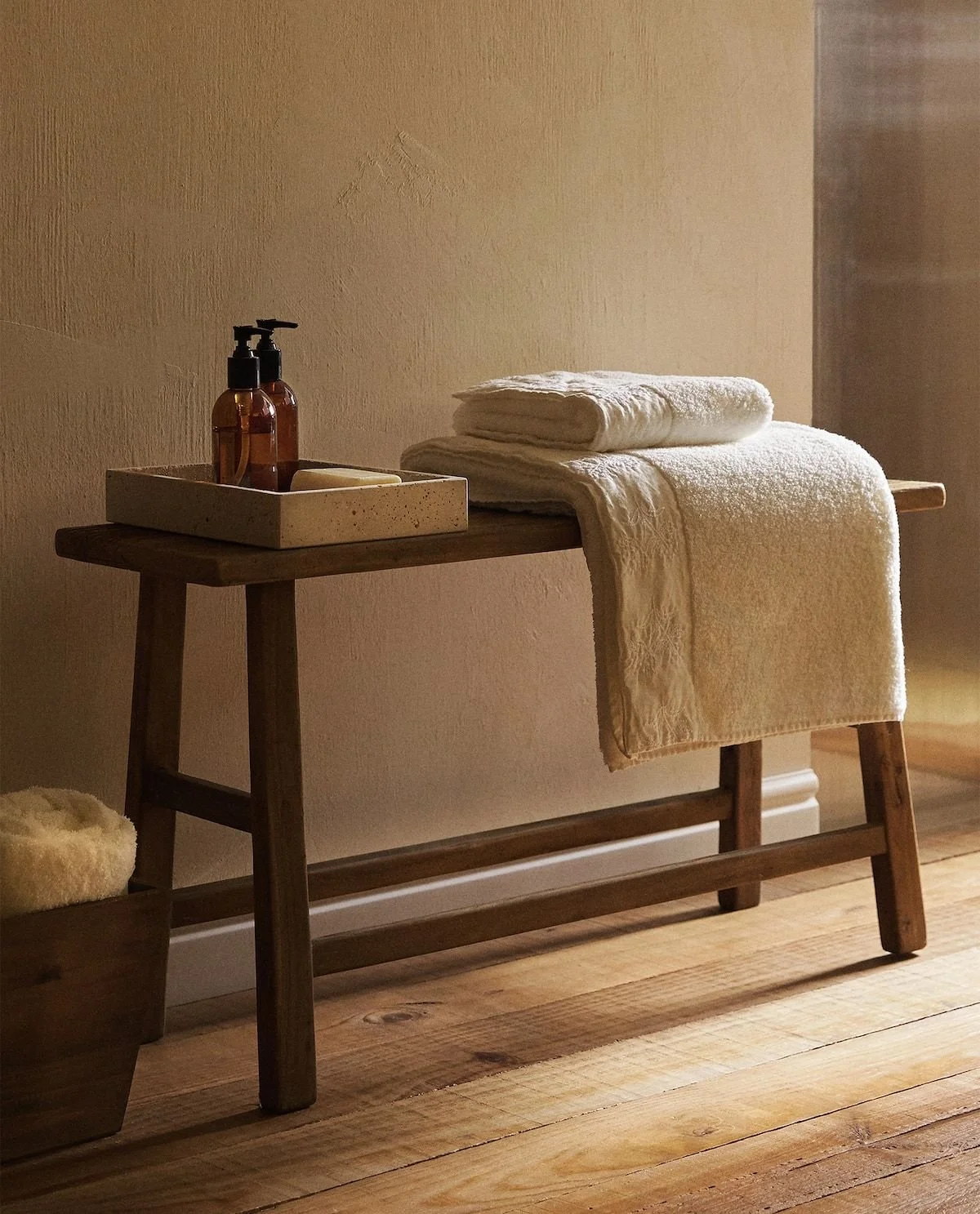 A wooden bench with folded white towels and a tray of soap and bottles, set against a neutral wall and wooden floor.