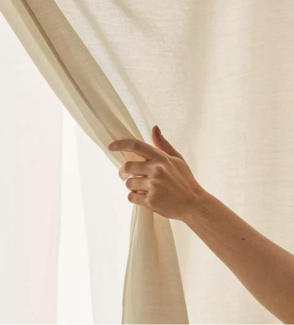 A person's hand pulling back a curtain in front of a window.
