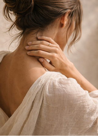 A woman with her hair tied back wearing a white off-the-shoulder blouse, gently touching her neck with one hand.