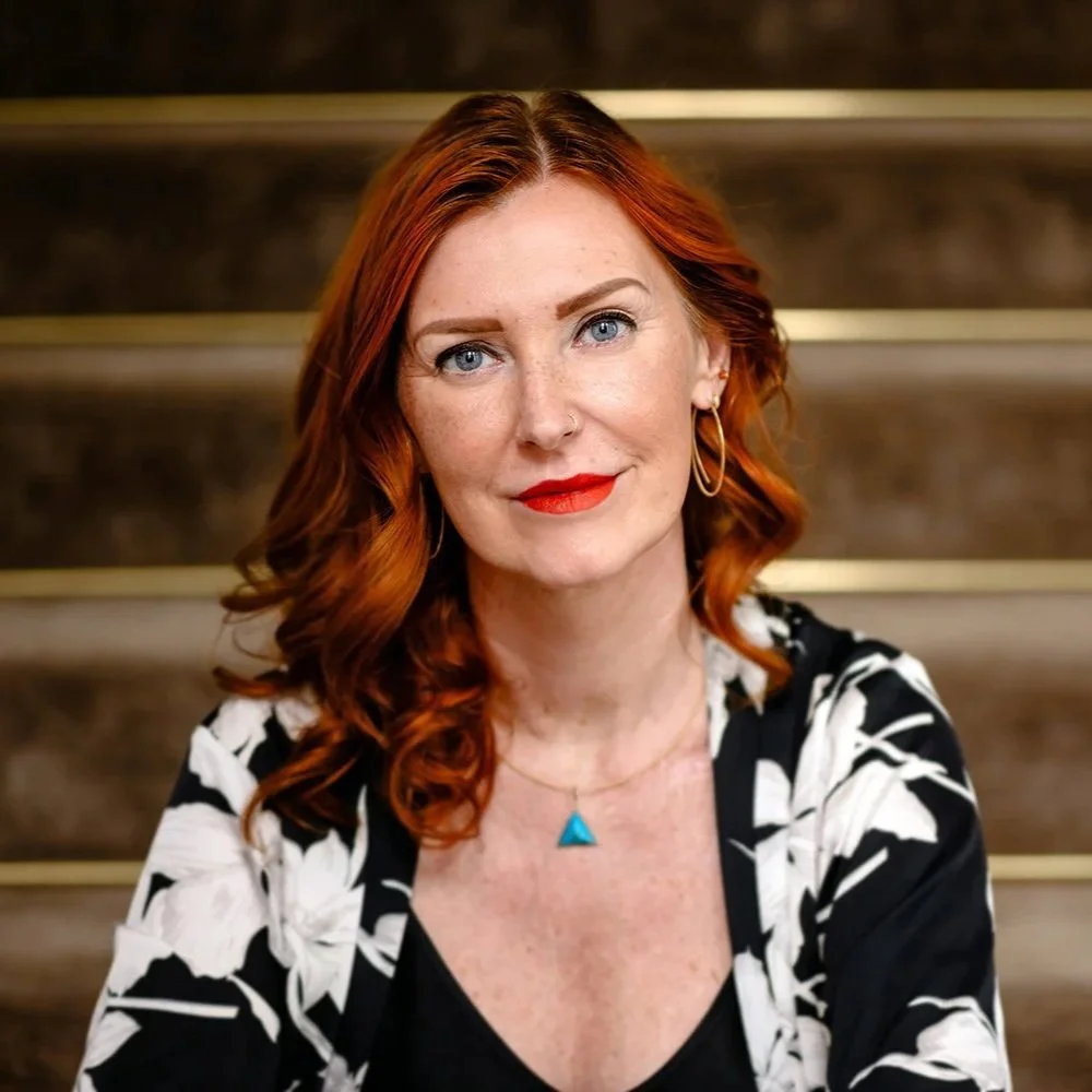 Porträt einer roten haare-d Frau, die vor einer Treppe sitzt, trägt ein schwarzes und weißes Musteroberteil, goldene Ohrringe und eine blaue Kette.