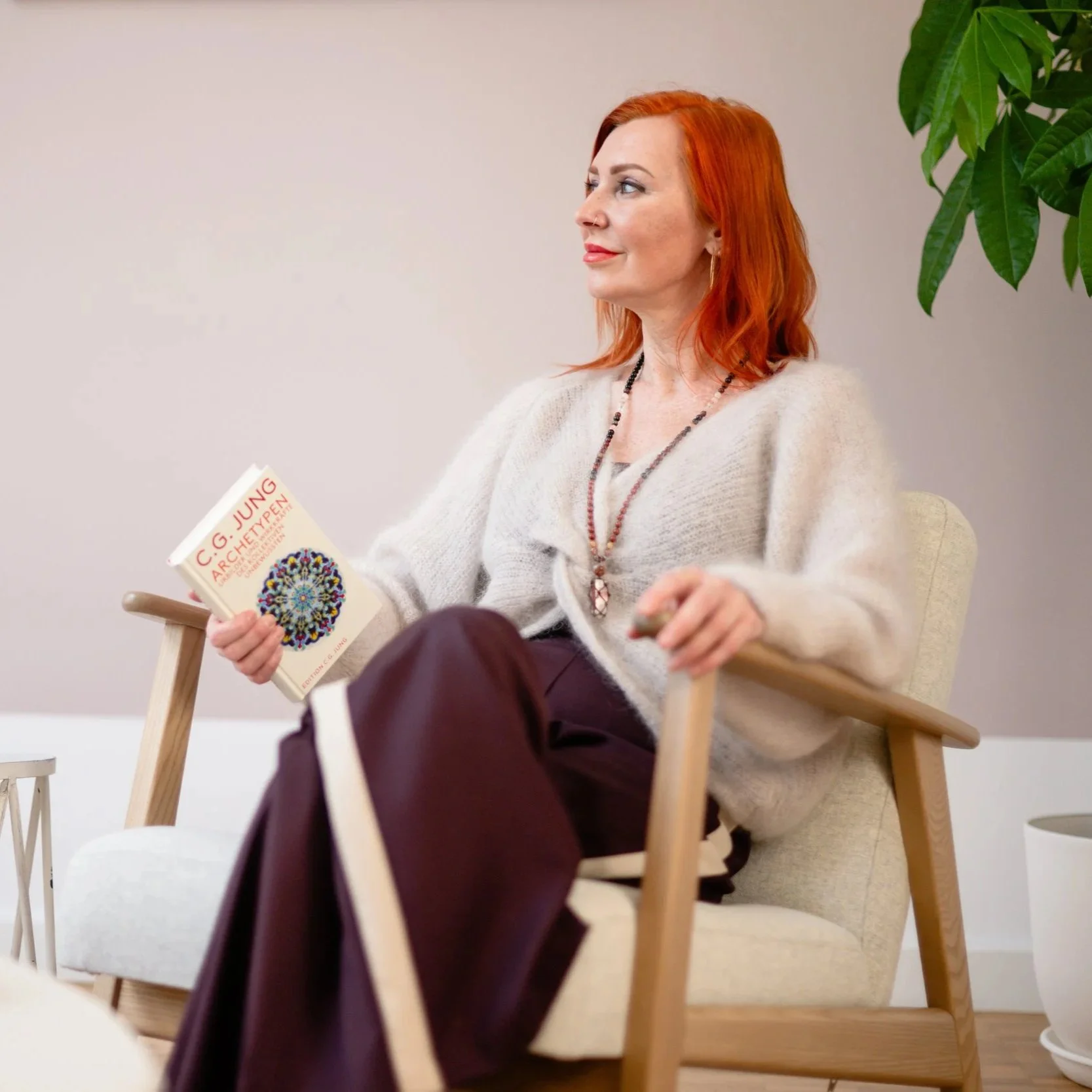 Eine Frau mit roten Haaren liest ein Buch in einem Wohnzimmer, sitzend in einem bequemen Stuhl.