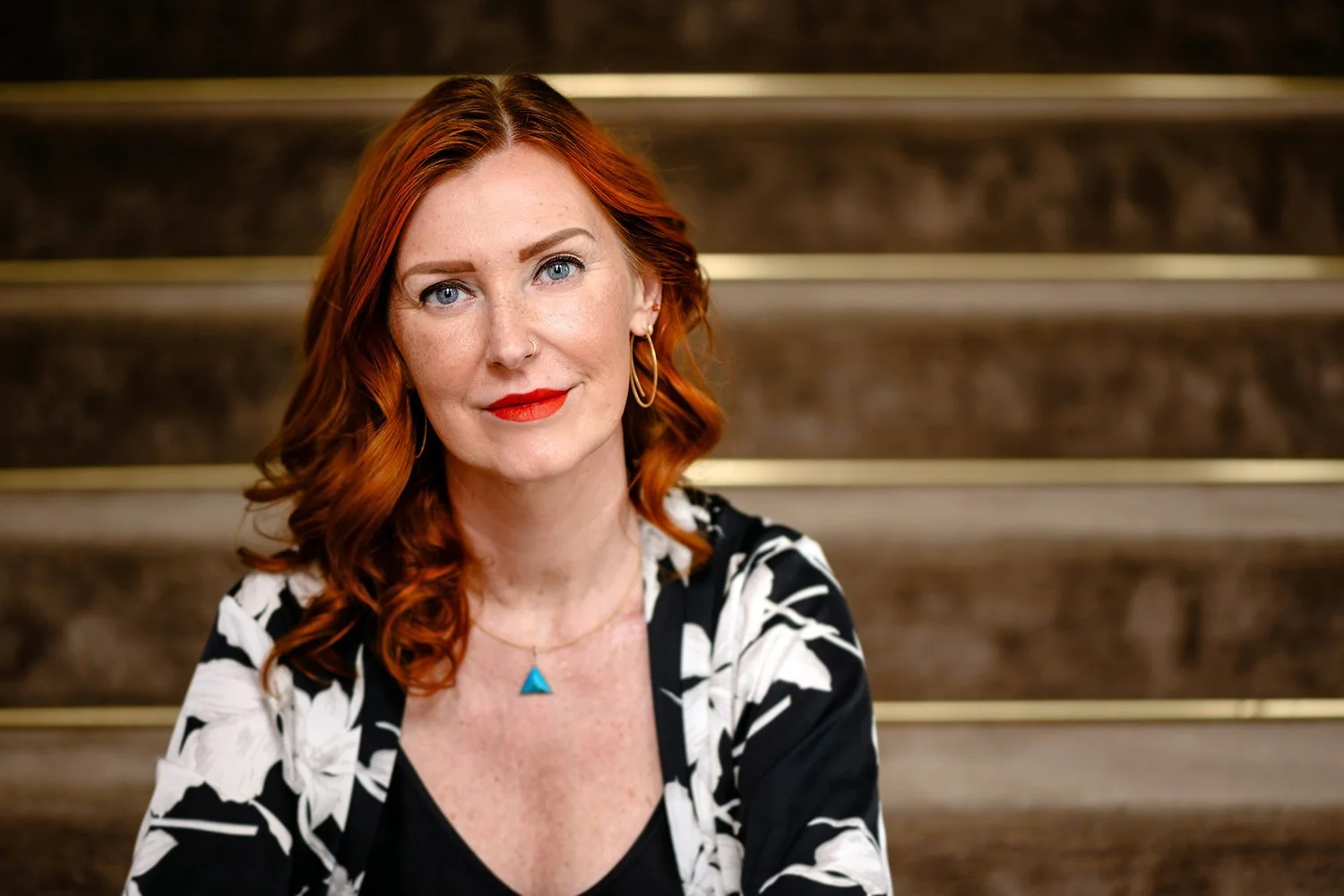 Eine Frau mit roten, lockigen Haaren, rotem Lippenstift, Ohrringen und einem blauen Halsreif sitzt auf einer Treppe aus Marmor.