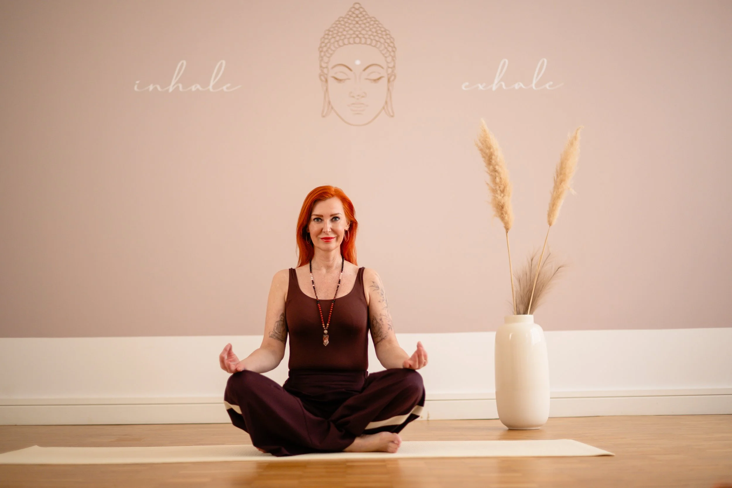 Eine Frau mit roten Haaren sitzt in einer Yoga-Haltung auf einem Matte im Raum, im Hintergrund eine Wand mit der Aufschrift 'inhale' und 'exhale' und einer stilisierten Gesichtsskizze. Eine weiße Vase mit Pampasgras steht rechts neben ihr.
