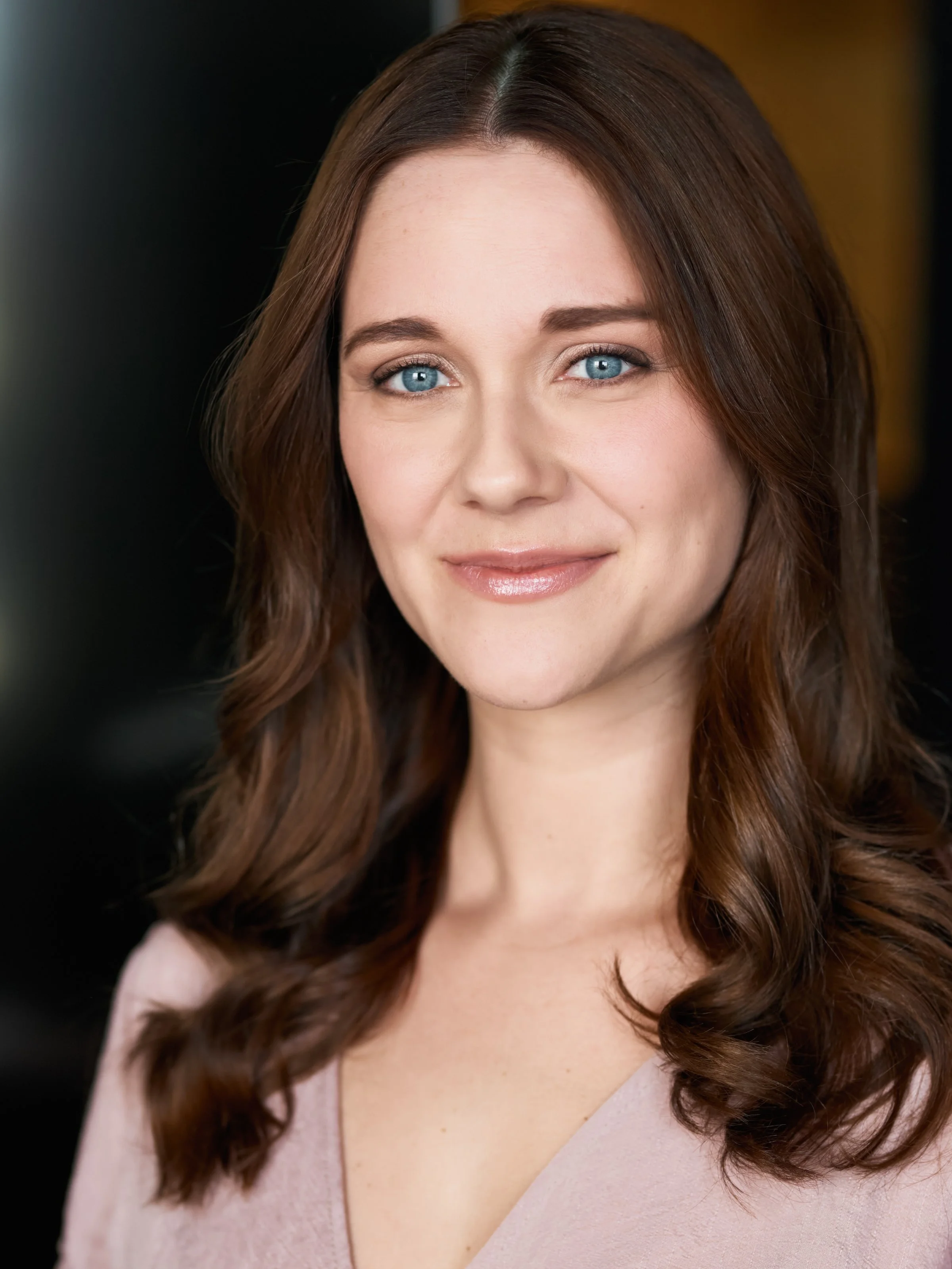 A woman with long, wavy brown hair and blue eyes, smiling softly at the camera.