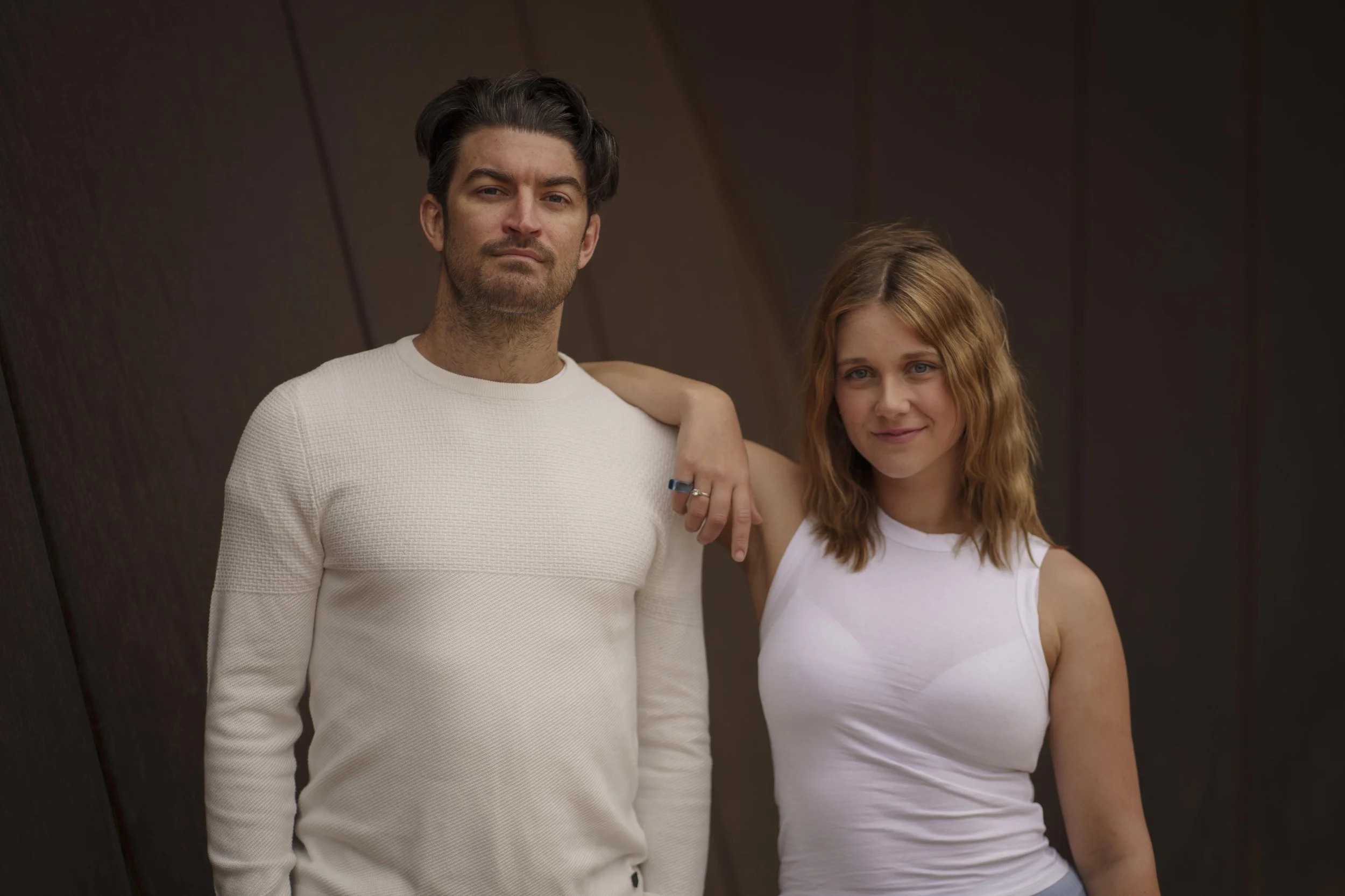 A man and a woman standing closely together with the man's arm around the woman's shoulder, both looking at the camera. The woman is wearing a white sleeveless top, and the man is wearing a white long-sleeve shirt. They are standing against a dark, angled background.