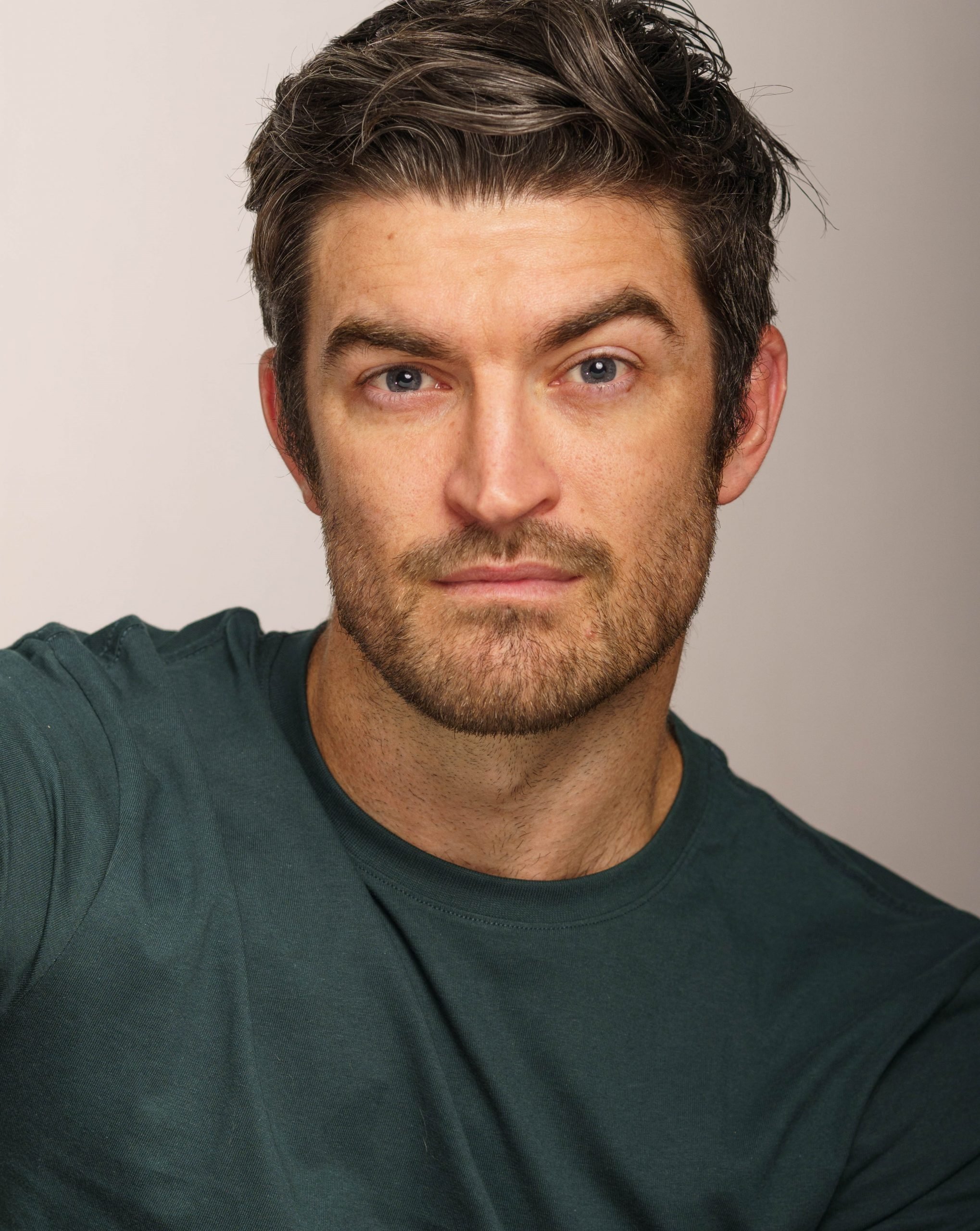 A young man with dark brown hair and a beard, wearing a dark green t-shirt, looking into the camera with a neutral expression.