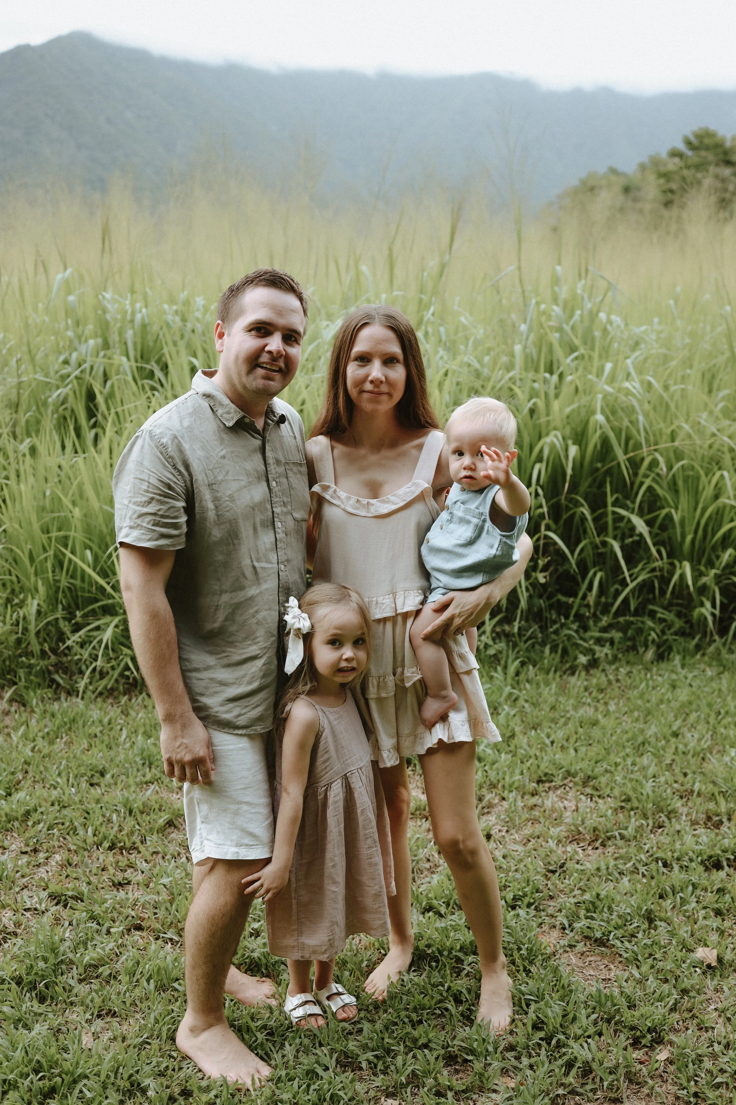 A family of five standing barefoot on grass, outdoors in a field with cairns sugarcane and mountains in the background. The family includes two adults, two young girls, and a toddler boy.