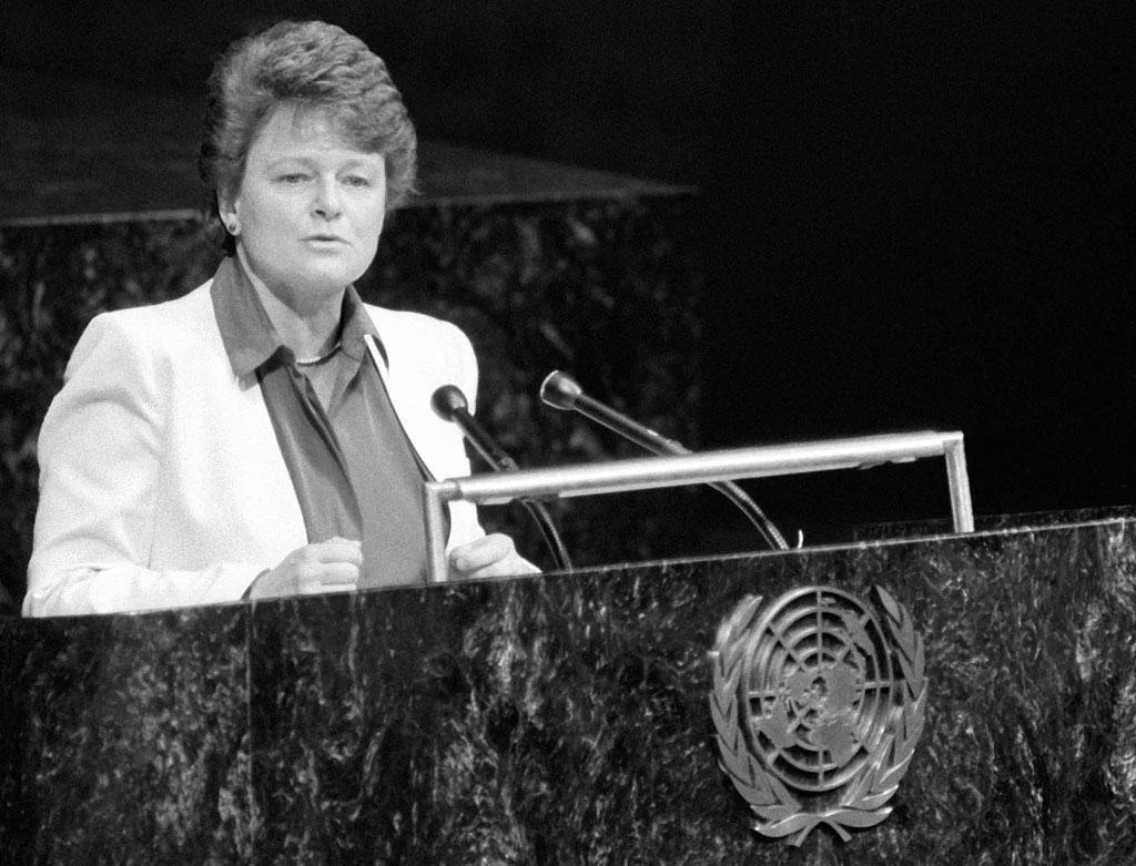 A woman standing at a podium with the United Nations emblem, speaking into microphones, in a formal setting.