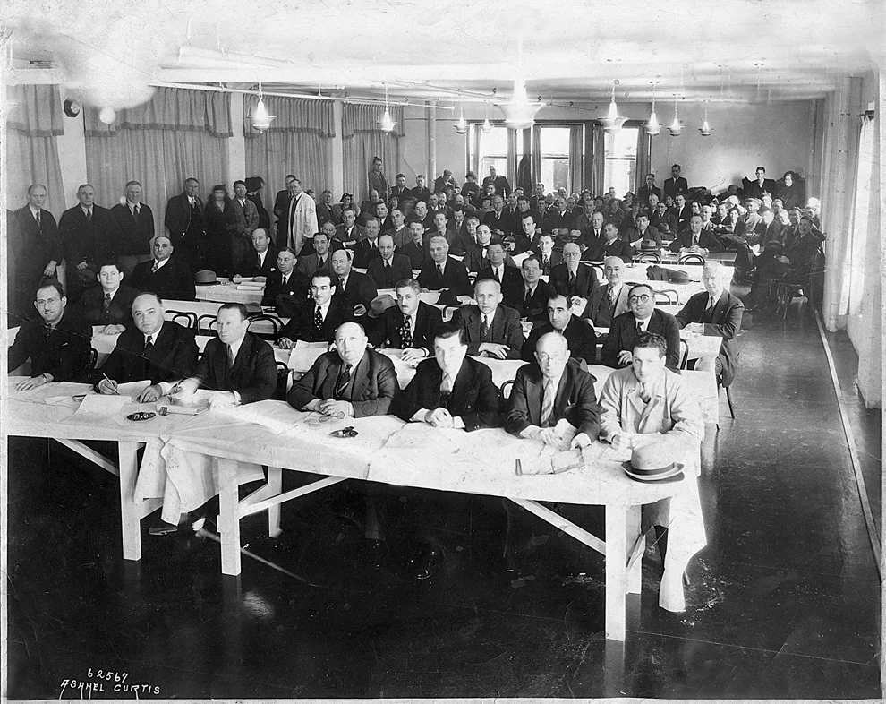A black and white photo of a large group of men seated at long tables in a conference or meeting room, with some standing at the back. The room has curtains and large windows, and the men are dressed in suits.