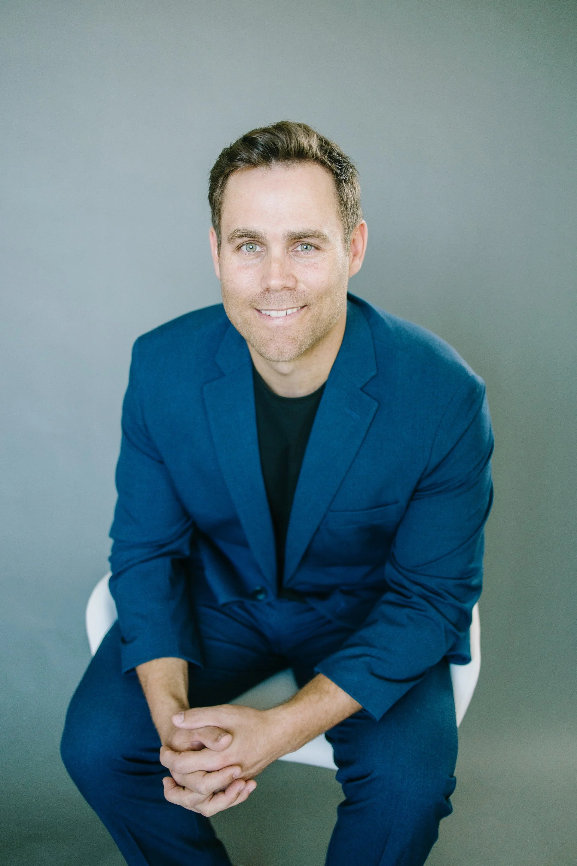 A smiling man with short brown hair and blue eyes wearing a blue blazer and black shirt, sitting on a white chair against a plain, gray background.