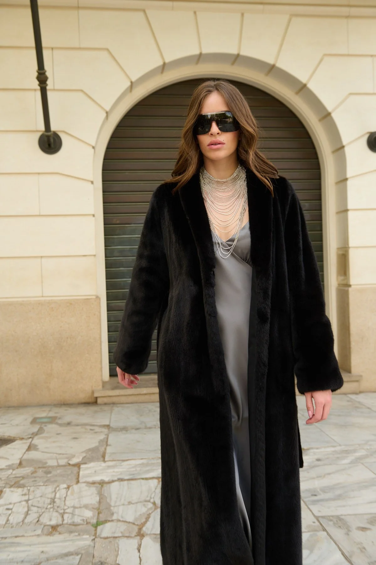 Woman walking outdoors in front of a building with an arched doorway, wearing large black sunglasses, a black fur coat, and layered silver necklaces.