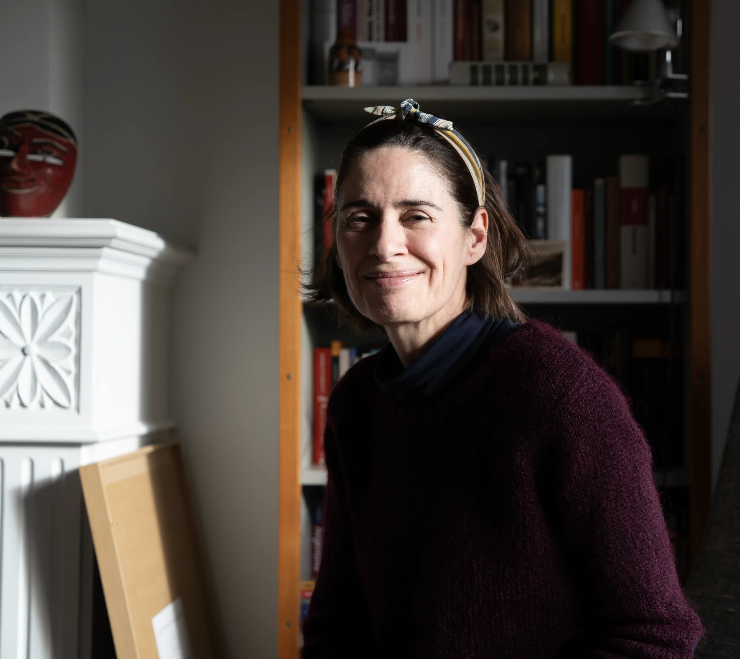 A woman with short brown hair wearing a headband, smiling in a room with bookshelves and a sculpture of a traditional mask.