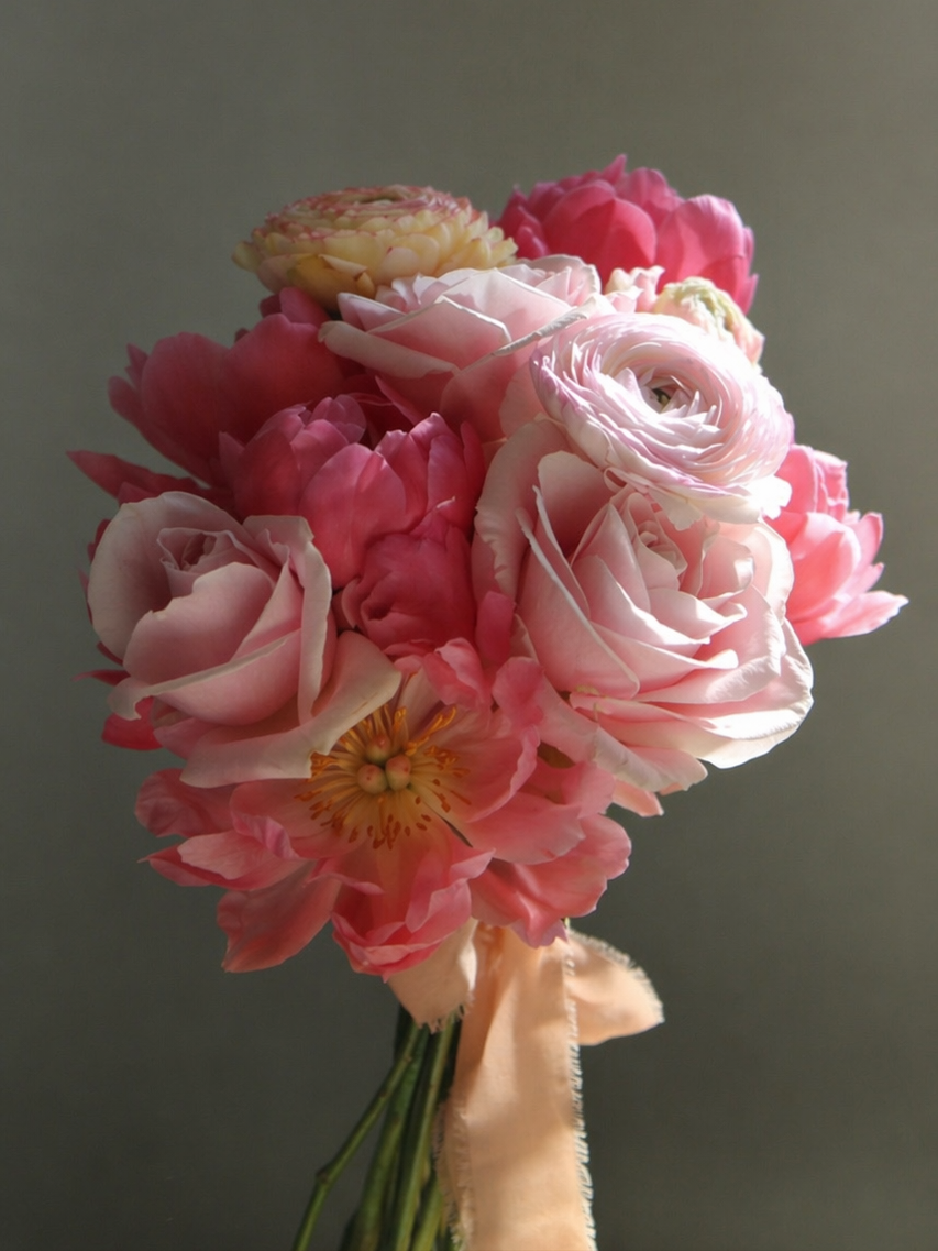 A delicate bridal bouquet featuring layers of blush pink ranunculus, soft pink roses, and creamy white peonies, finished with a long, trailing peach silk ribbon.