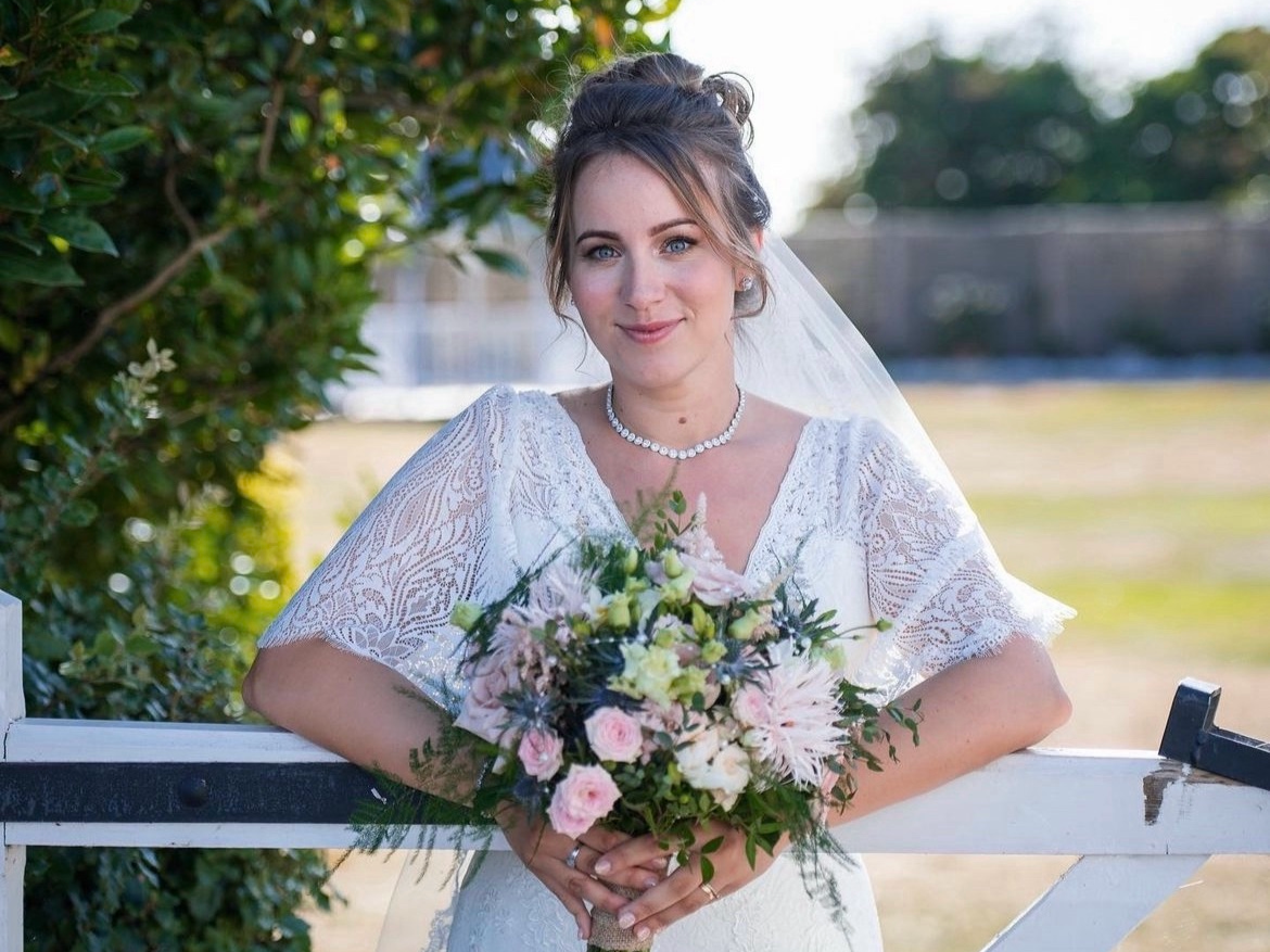 A romantic portrait of a bride leaning on a white wooden gate, holding a textural garden-style bouquet of soft pink spray roses, dahlias, and blue thistle with wild greenery.