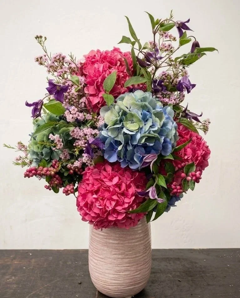 A lush floral arrangement in a textured pink ceramic vase, featuring hot pink and blue hydrangeas, purple clematis vines, and pink waxflower accents against a neutral background.