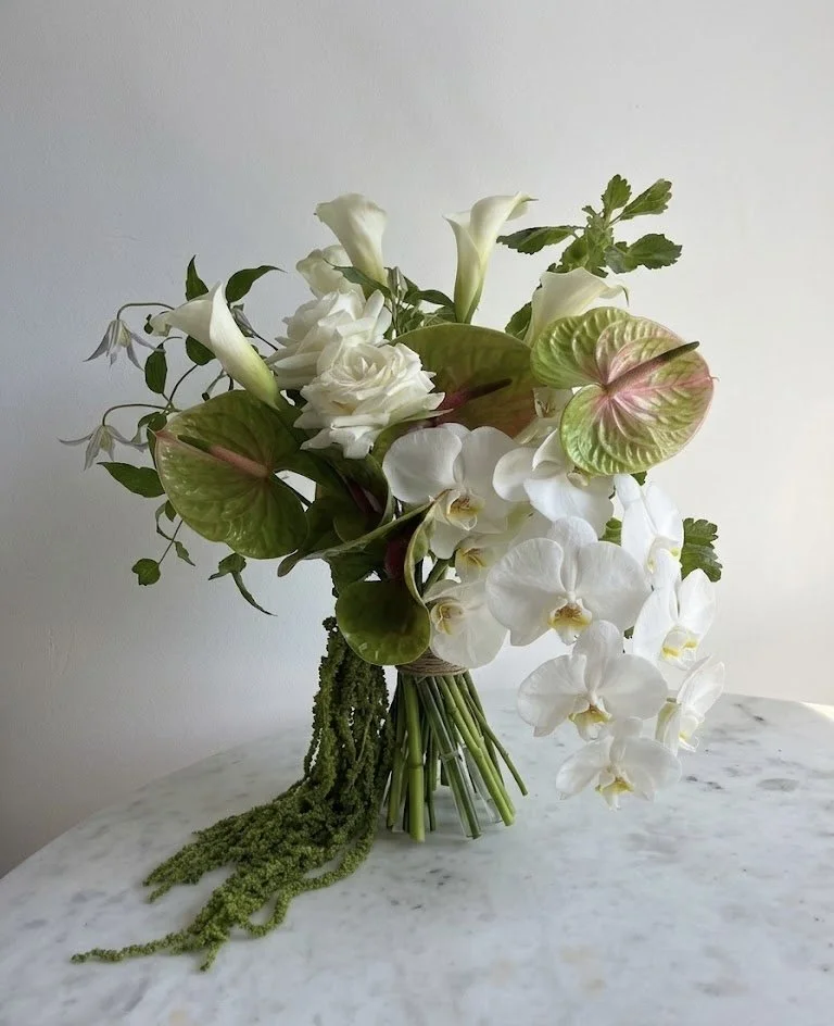 A modern, sculptural bridal bouquet featuring cascading white Phalaenopsis orchids, green anthurium with pink veins, white calla lilies, and trailing green amaranthus on a marble surface.