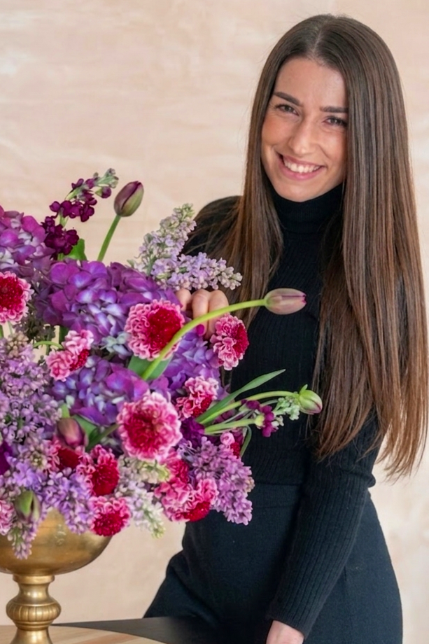 A woman with long brown hair, wearing a black turtleneck, smiling and holding a large colorful bouquet of purple, pink, and red flowers in a gold vase.