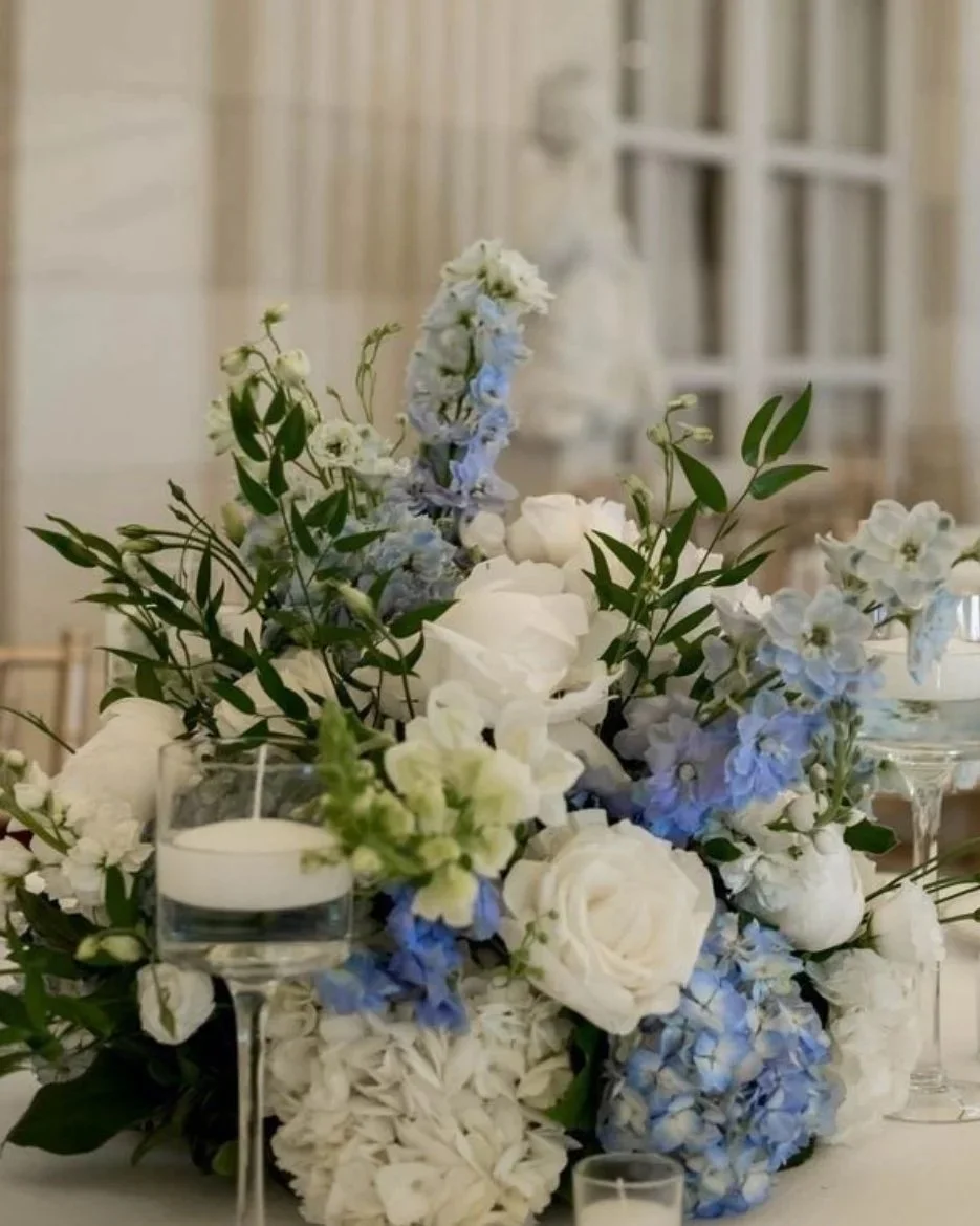 Elegant flower arrangement with white roses, blue hydrangeas, and green leaves, accompanied by white candles in glass holders, set on a table in a bright room.