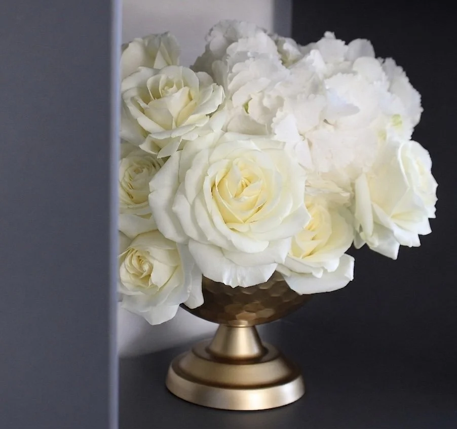 A classic, monochromatic masterpiece of full-bodied white roses nestled in a gold hammered compote. This arrangement plays with soft light and rich textures to create a "refined floral reality" for any luxury event.