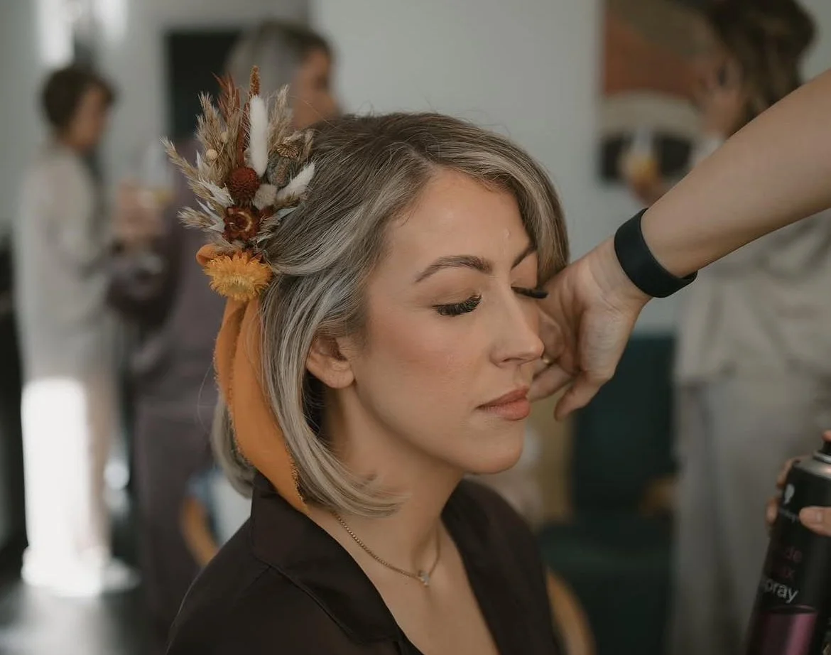 A bride during wedding preparations wearing a bespoke hair accessory made of dried textures, including bunny tails and strawflowers in terracotta and ochre tones, finished with a silk ribbon.