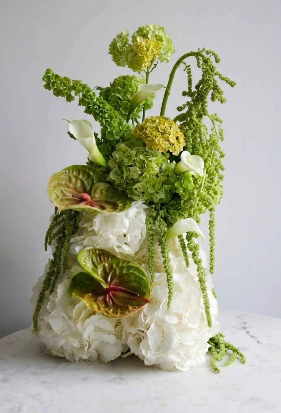 A unique, sculptural floral installation featuring a dense base of white hydrangeas, accented with green anthurium, sleek white calla lilies, and cascading green amaranthus on a marble tabletop.