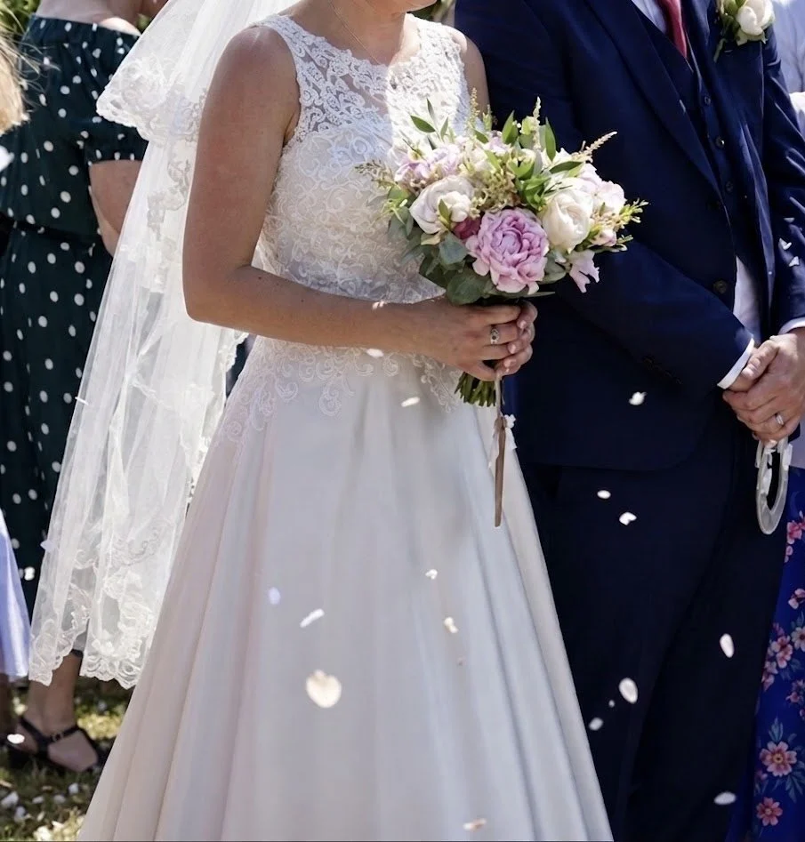 A bride in a lace-topped wedding dress holding a romantic garden-style bouquet featuring pink peonies, ivory roses, and textural greenery during an outdoor wedding ceremony.