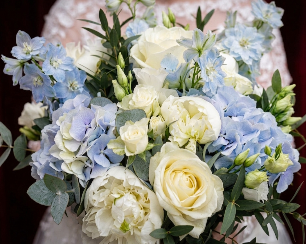 A close-up of a full bridal bouquet featuring large white roses, creamy peonies, light blue hydrangeas, and pale blue delphinium stems, accented with eucalyptus and green foliage.