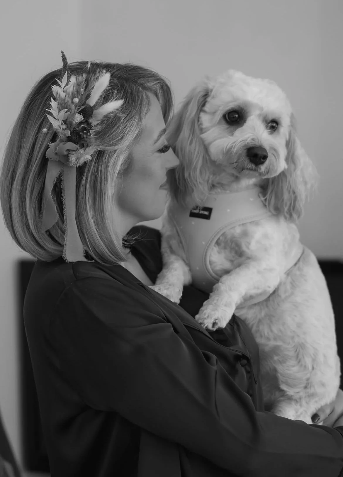 A black and white bridal portrait of a bride wearing a textural dried flower hairpiece with silk ribbons, holding her dog before the wedding ceremony.