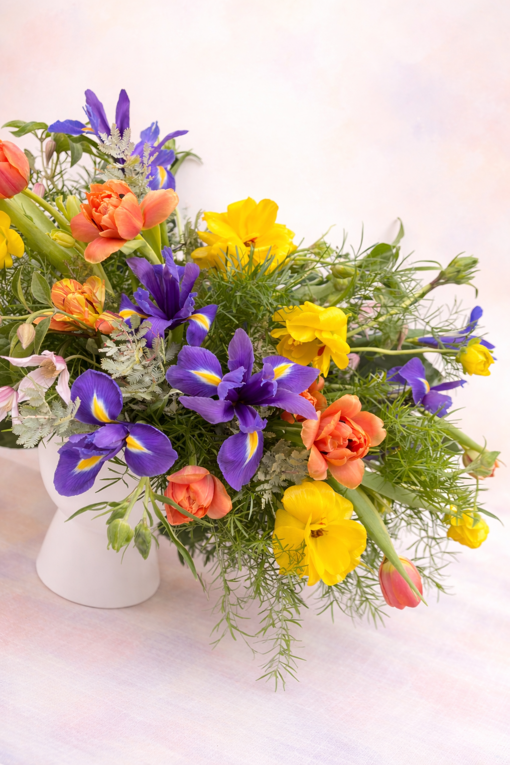 A bright and airy seasonal floral arrangement in a modern white ceramic vase, featuring bold purple irises, orange double tulips, and yellow ranunculus accented with feathery green foliage.