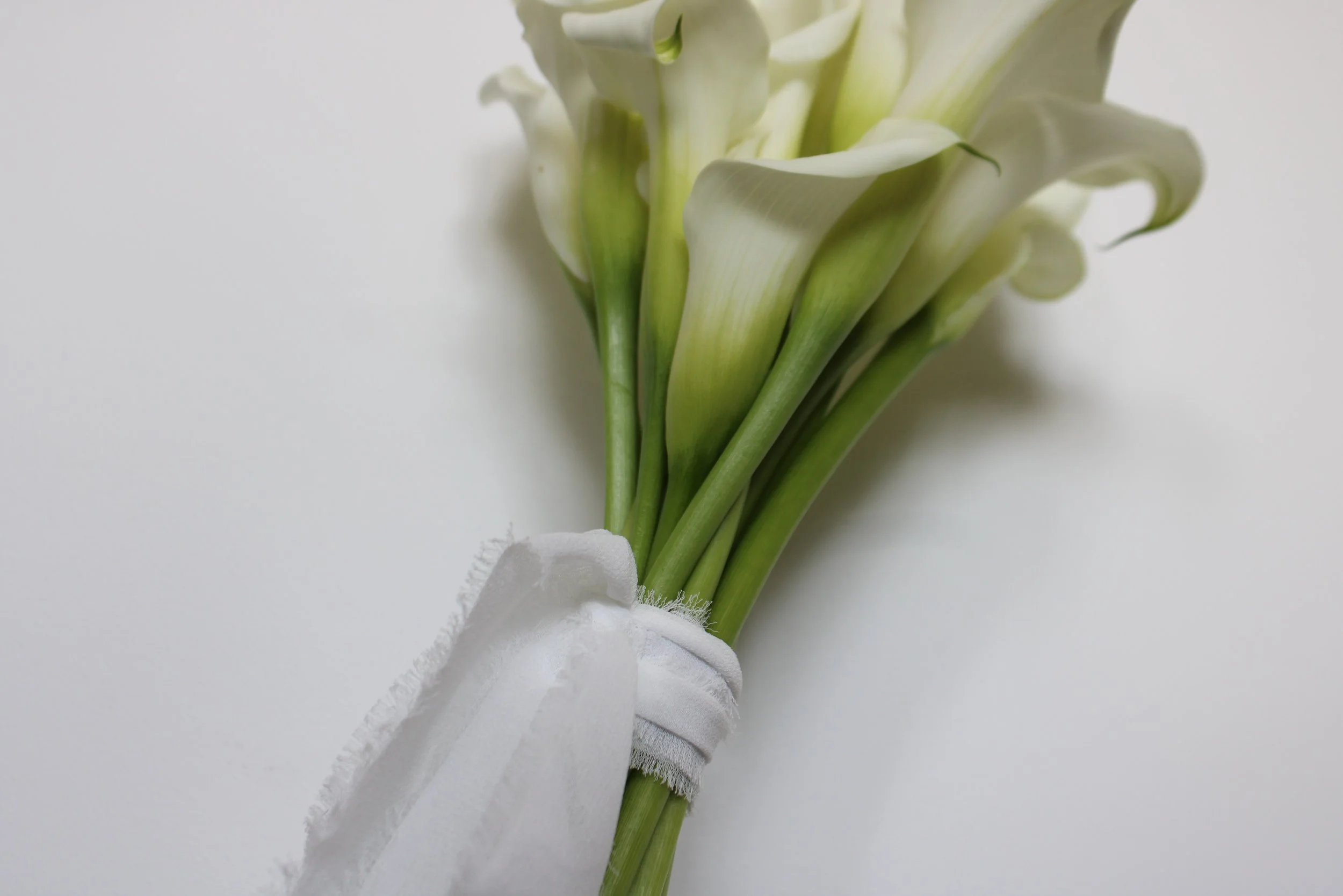 A minimalist bridal bouquet of crisp white calla lilies with vibrant green stems, bound tightly with a textured, frayed-edge white silk ribbon against a clean white background.