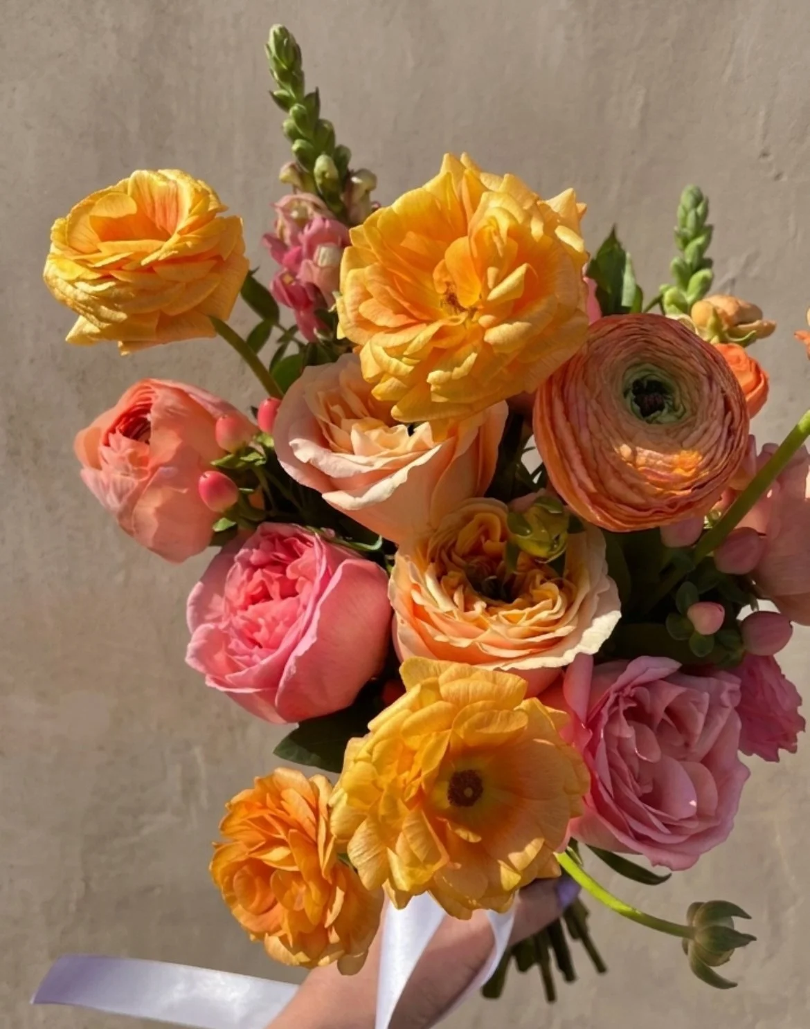 Warmth, light, and garden-inspired romance. This bespoke bridal bouquet is a curated mix of honey-toned butterfly ranunculus, peach garden roses, and textural accents. Tied with a signature flowing silk ribbon, it represents our dedication to creatin