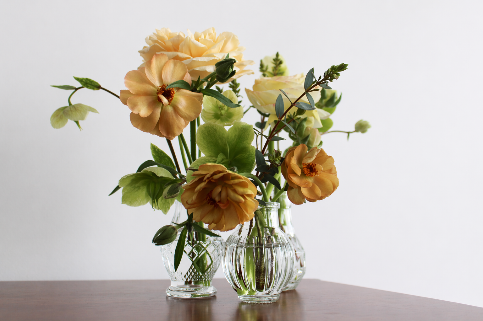 A delicate, garden-style floral arrangement in three small crystal and ribbed glass bud vases, featuring honey-colored butterfly ranunculus, soft peach roses, and green hellebores on a dark wood surface.