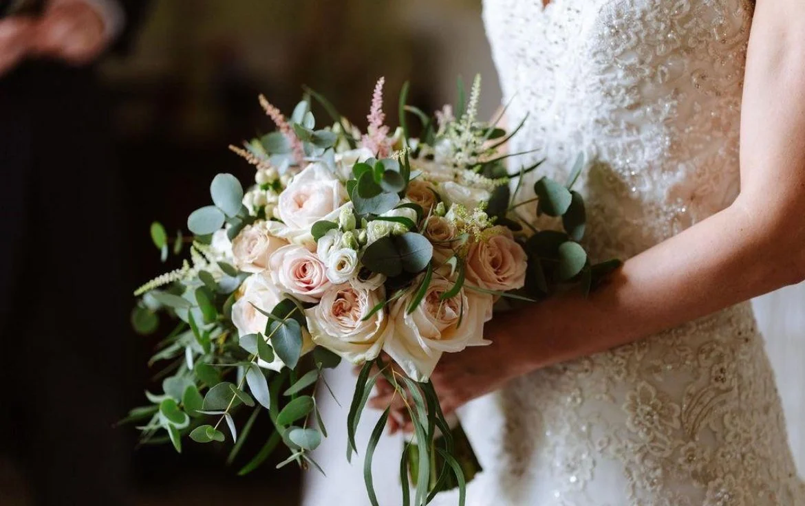 A bride in a detailed lace wedding gown holding a romantic, organic-style bouquet of blush garden roses, white lisianthus, and pink astilbe, accented with trailing seeded and silver dollar eucalyptus.