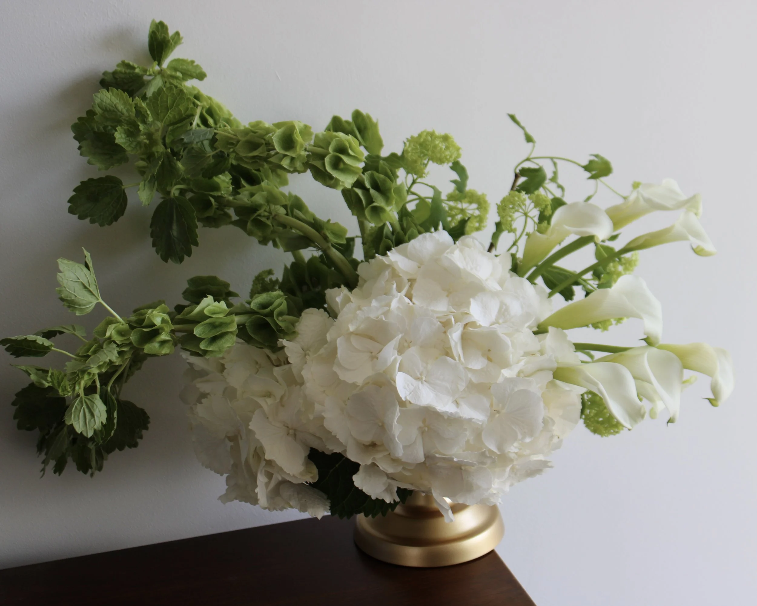 A masterclass in form and movement. This modern, asymmetrical centerpiece balances the soft volume of white hydrangeas with the sleek, sweeping lines of calla lilies and architectural greenery. Set in a classic gold compote, it is a sophisticated sta