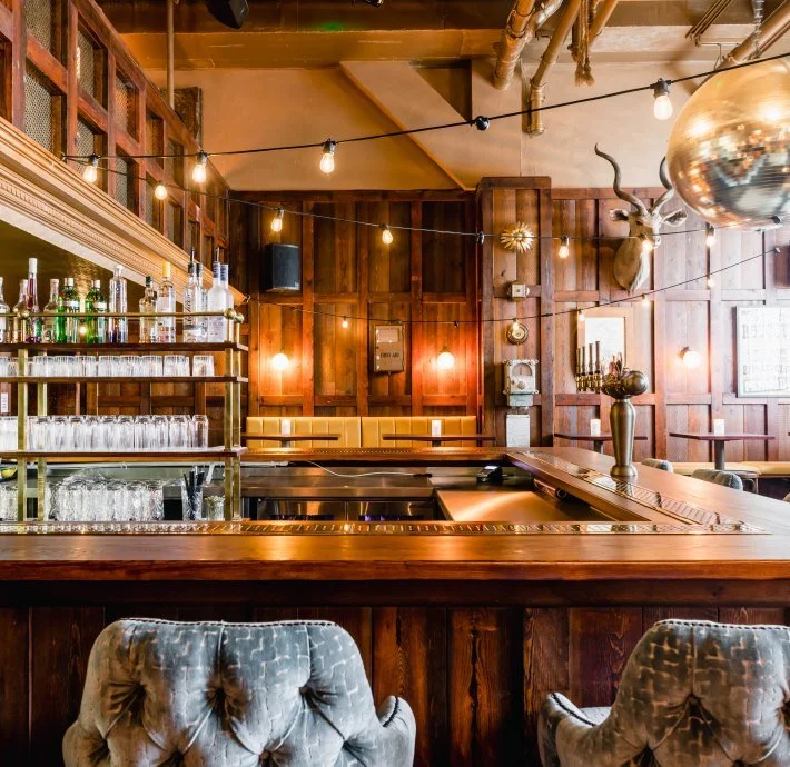 Interior of a secret bar with wooden walls, a bar counter with glasses and bottles, vintage seating, and warm lighting including hanging bulbs and wall lamps, with hanging string lights and a mounted deer head with antlers.