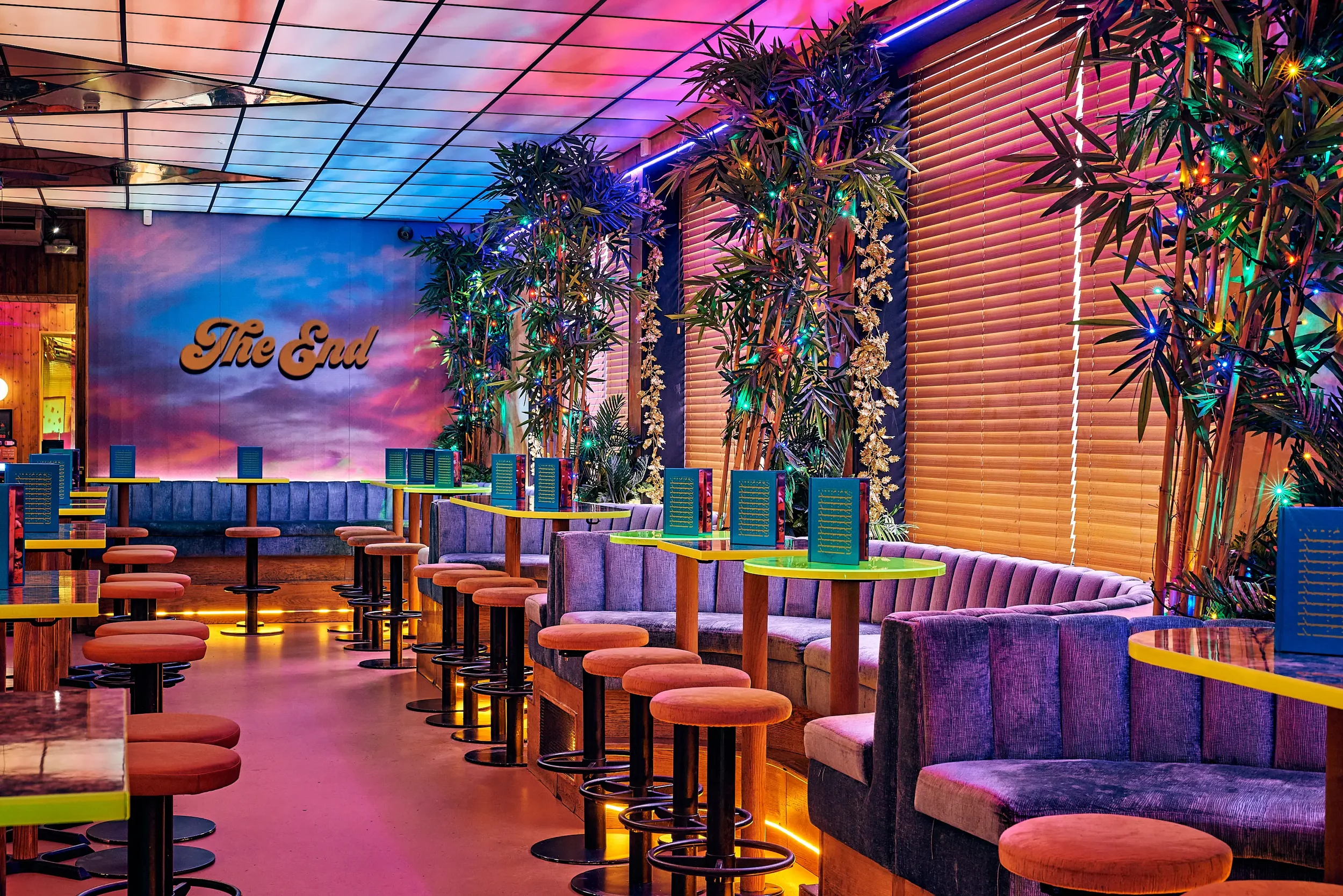Colorful cocktail bar, wooden tables, and a wall with "The End" sign, decorated with tropical plants and multicolored string lights.