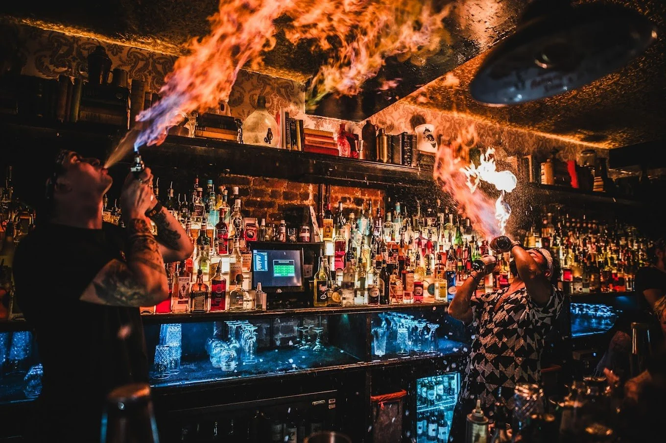 Two bartenders in a bar, each exhaling fire while performing a fire-breathing trick. The background is filled with liquor bottles and bar equipment.