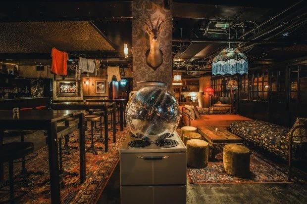 Interior of a cozy, eclectic lounge or bar with vintage decor. There is a disco ball on a white cabinet stove, a mounted deer head on a central pillar, and various seating areas with mismatched furniture and rugs. Clothes are hanging on a line in the