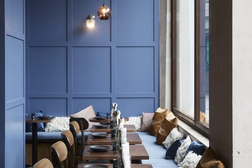 A cozy restaurant corner with a blue paneled wall, wooden tables set with black plates, and a bench with pillows by a large window.