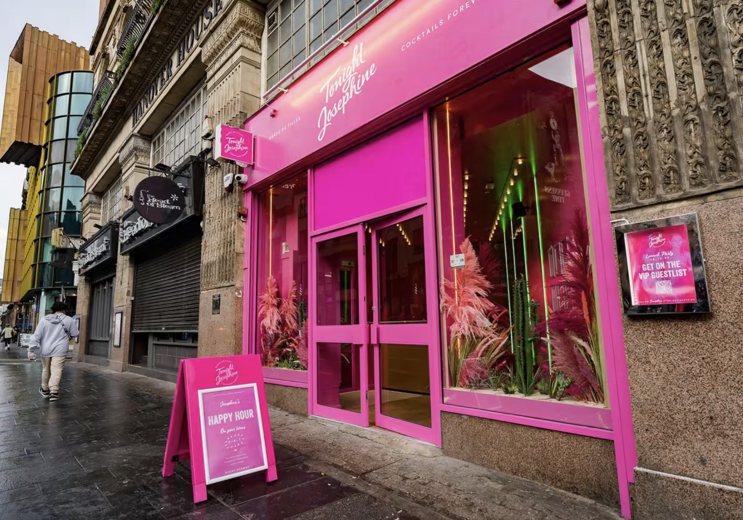 Pink storefront for a bar called 'Tonight Josephine' with plants inside, pink signboard, and sidewalk signboard advertising happy hour, on a city street with pedestrians and neighboring buildings.