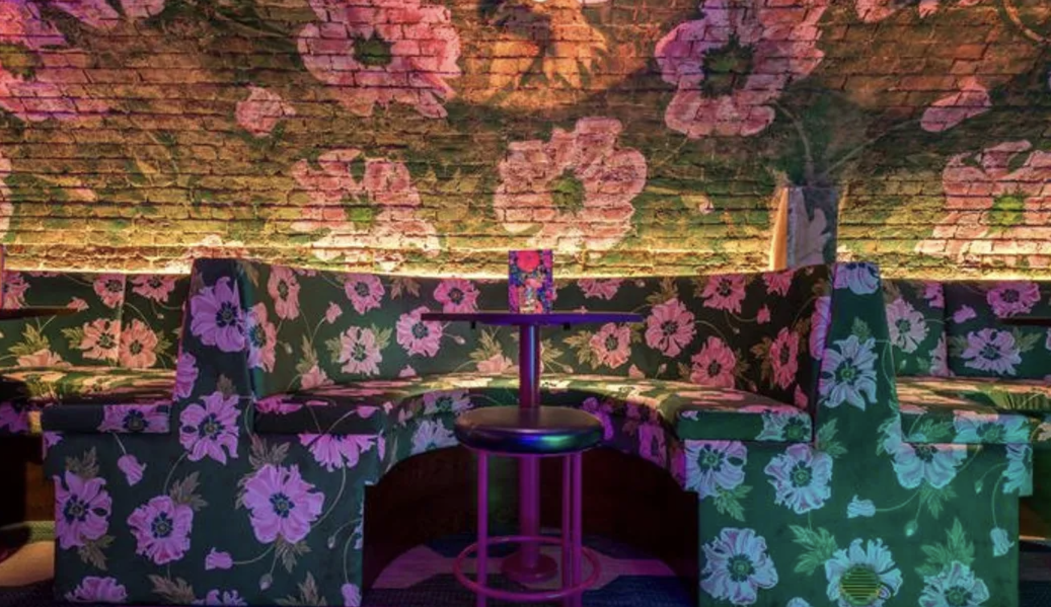 Colorful, floral-patterned seating arrangement with a small round table and a decorative glass on top, set against a vibrant, flower-themed brick wall backdrop in a cozy, ambient space.