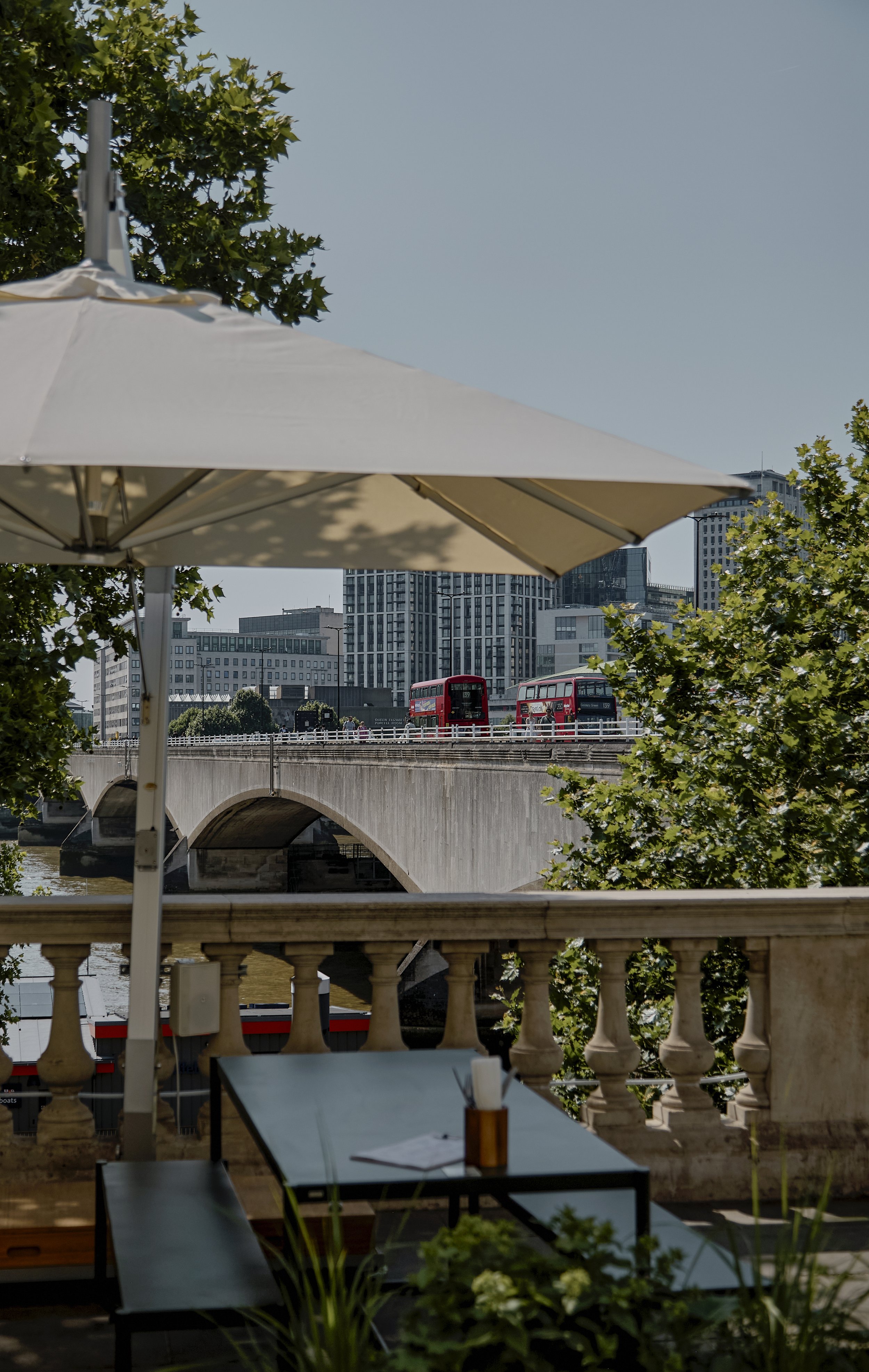 A view from a patio or balcony overlooking a bridge with two red double-decker buses crossing, surrounded by modern high-rise buildings, framed by a large white umbrella and green trees in the foreground.
