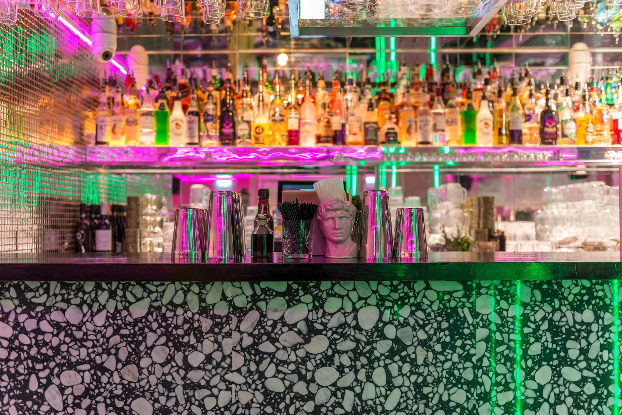 Bar with colorful bottles of alcohol, glasses hanging upside down, neon lights, and decorative items including a pink bust and black straw containers.