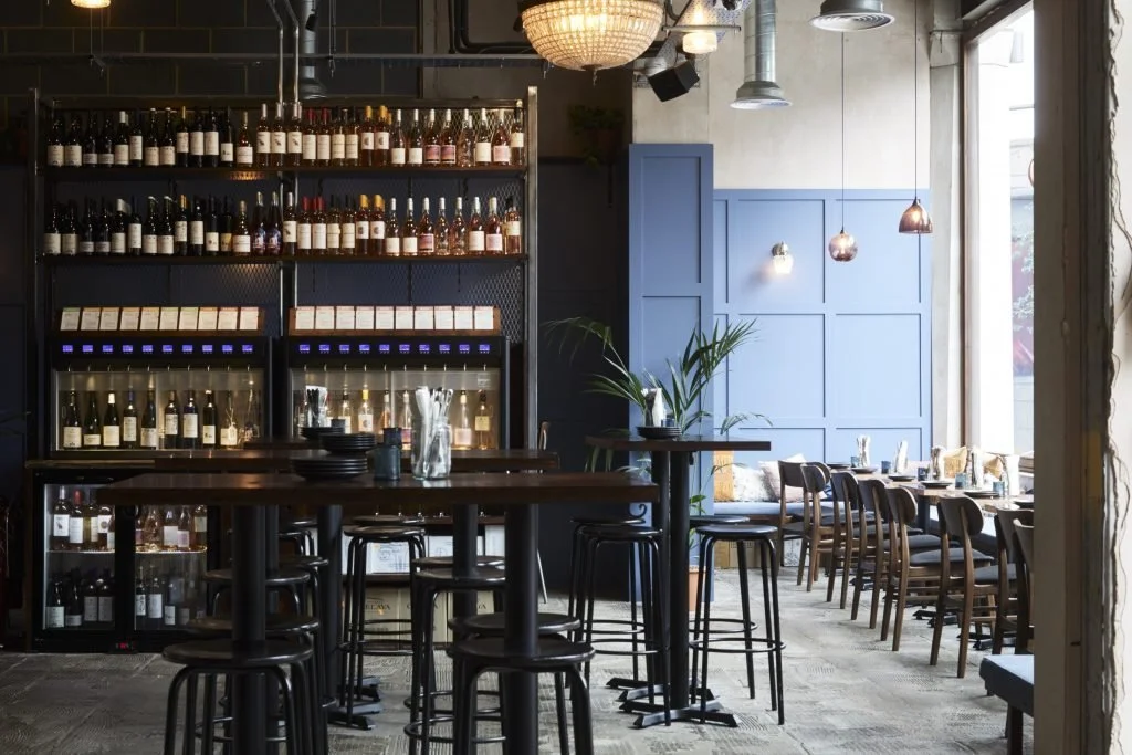 Interior of a modern wine bar with a wall of wine bottles, bar tables with stools, and a seating area near large windows with natural light.