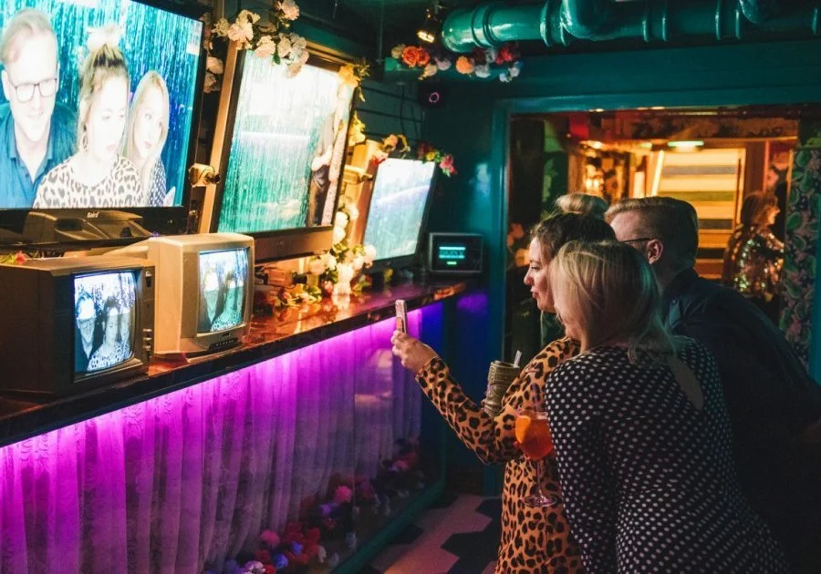 People watching vintage TVs in a colorful, decorated room, some taking photos with a smartphone.