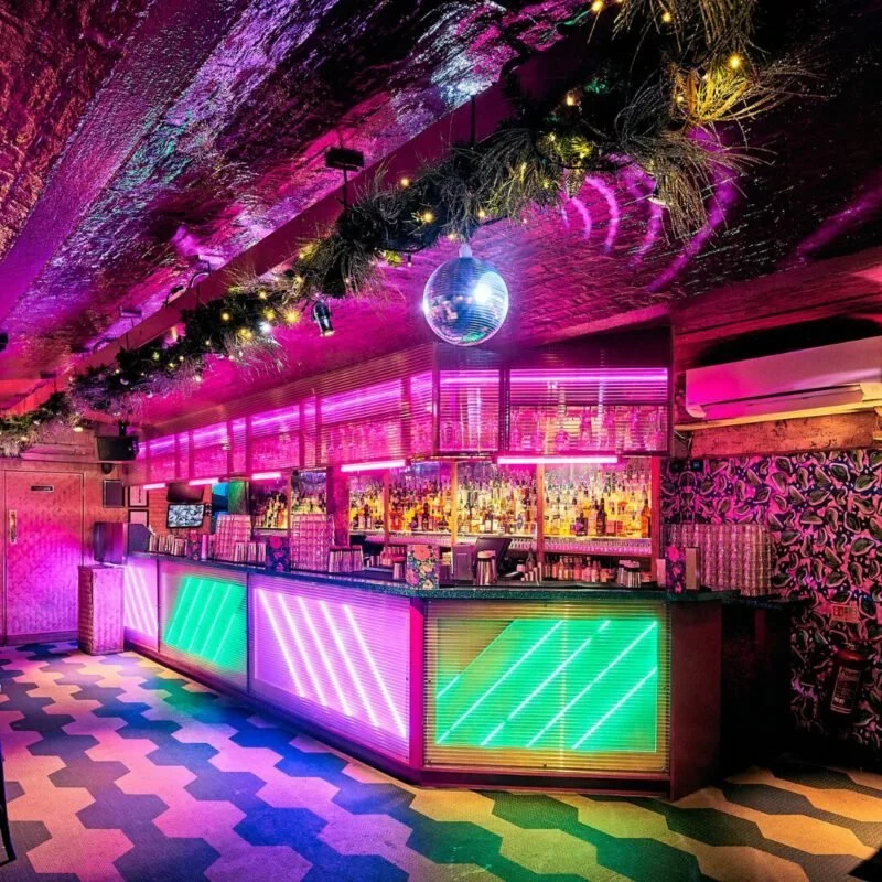 A vibrant nightclub bar with neon pink, green, and purple lighting, a glittering disco ball hanging from the ceiling, and festive greenery and lights decorating the ceiling.